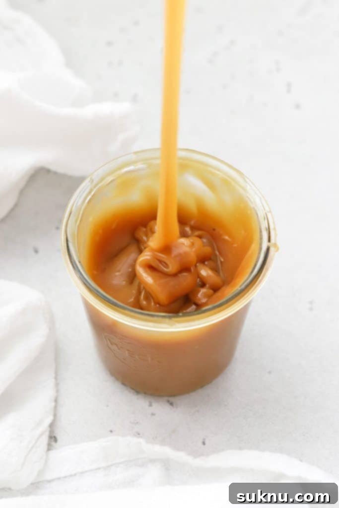Pouring rich, thick homemade caramel sauce into a clear glass jar, highlighting its luscious texture.