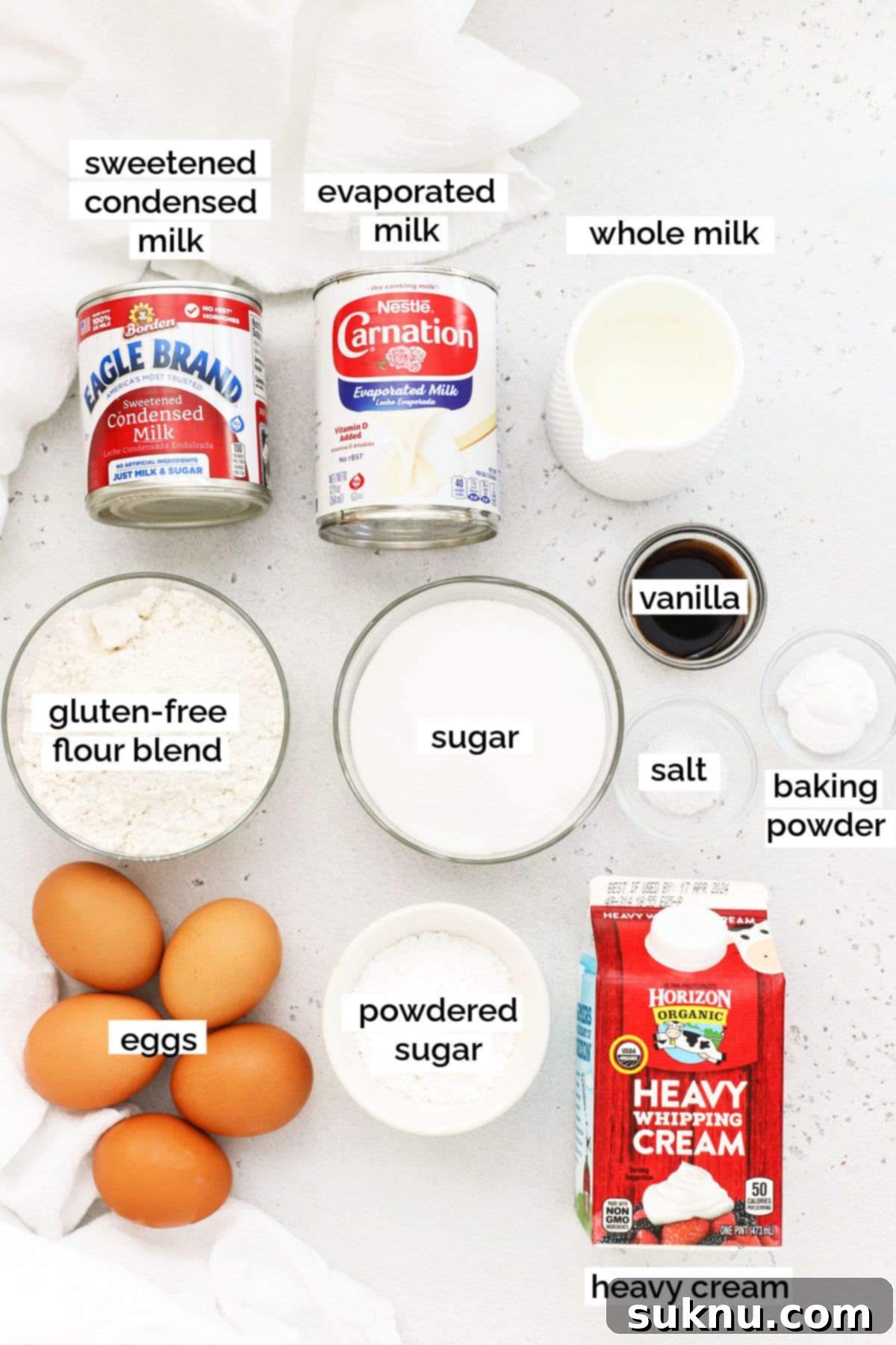 An array of fresh ingredients for baking gluten-free tres leches cake, including flour, eggs, and cans of milk.