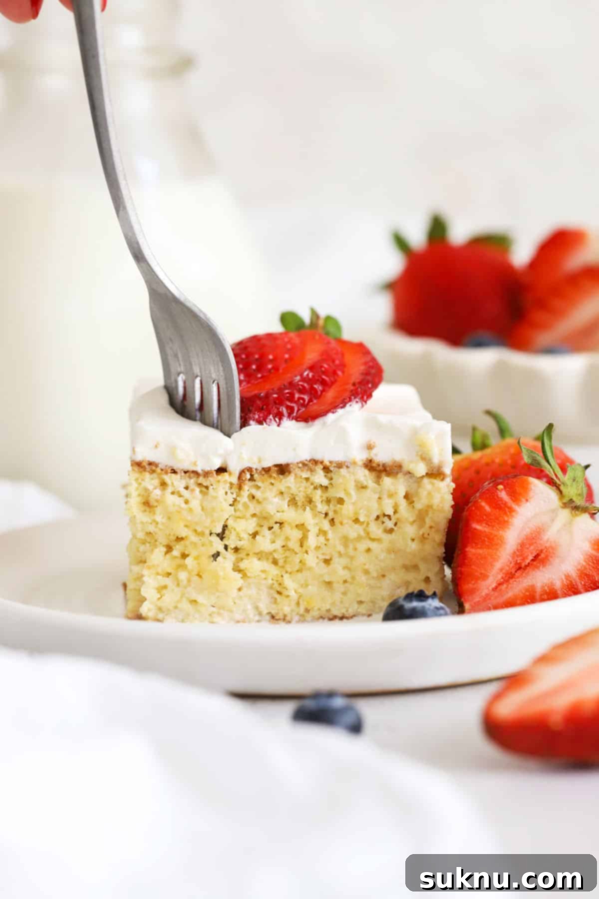 A fork taking a delicious bite from a slice of gluten-free tres leches cake, showing its moist texture and berry topping.