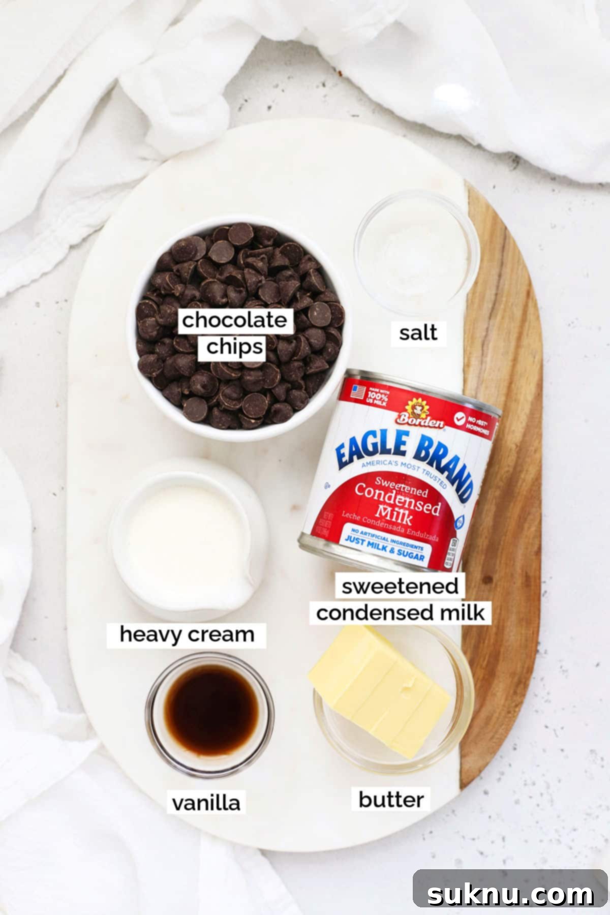 A collection of simple ingredients laid out for homemade hot fudge sauce: sweetened condensed milk, heavy cream, butter, dark chocolate, vanilla extract, and a pinch of salt.
