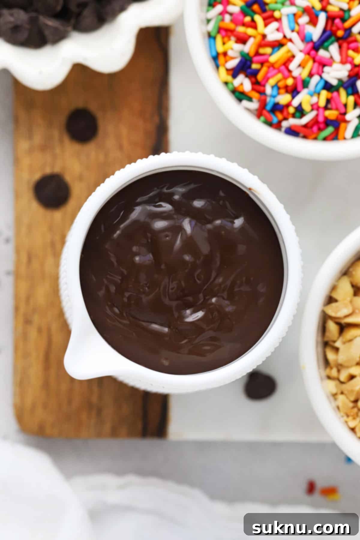 Old fashioned hot fudge sauce in a small white pitcher, surrounded by other ice cream toppings like sprinkles and nuts.
