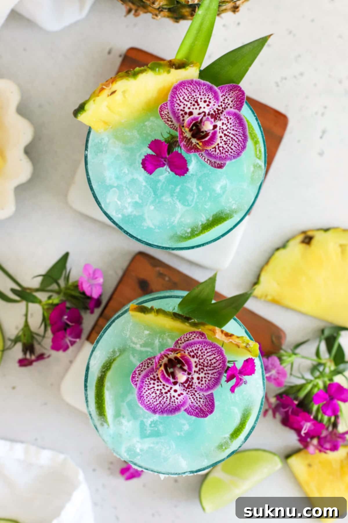 two blue hawaiian mocktails garnished with lime slices, fresh pineapple, and purple flowers