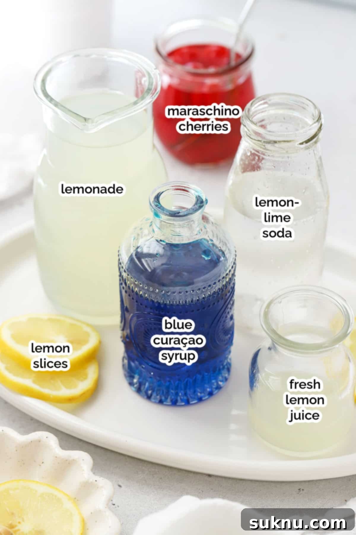 Azure Lagoon Cooler 3 Close-up of fresh ingredients for a Blue Lagoon mocktail: a bottle of blue curaçao syrup, fresh lemons, a pitcher of lemonade, and a clear glass filled with ice.