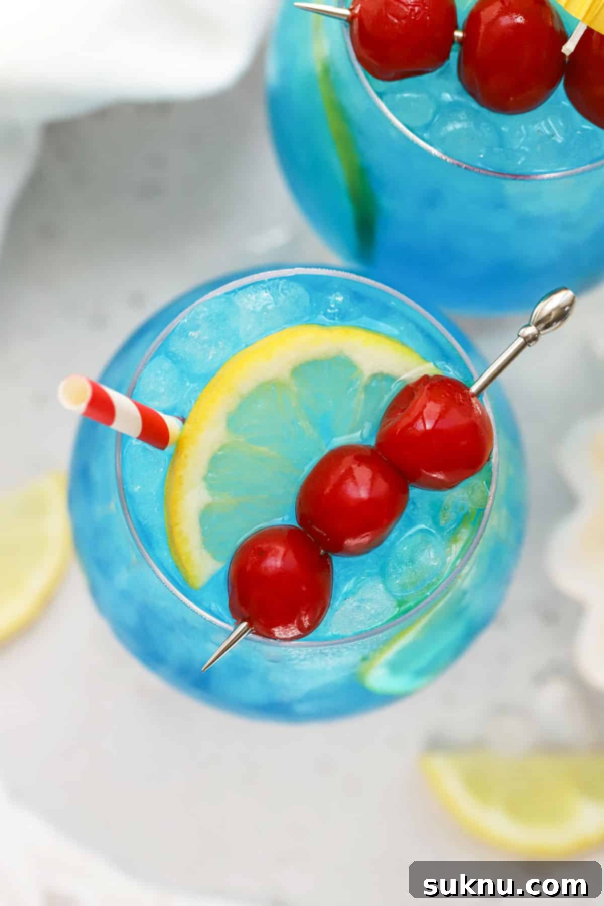Azure Lagoon Cooler 5 A beautifully garnished Blue Lagoon mocktail in a hurricane glass, adorned with lemon slices and maraschino cherries, ready to be enjoyed.