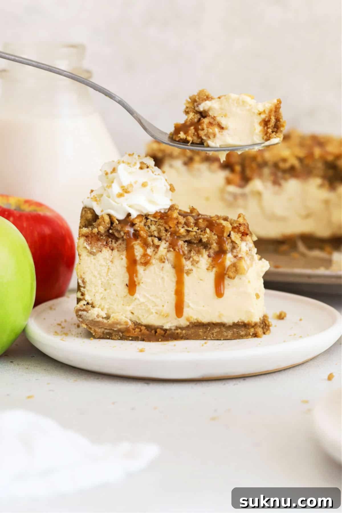 A fork taking a bite from a slice of gluten-free caramel apple crumble cheesecake on a white plate.