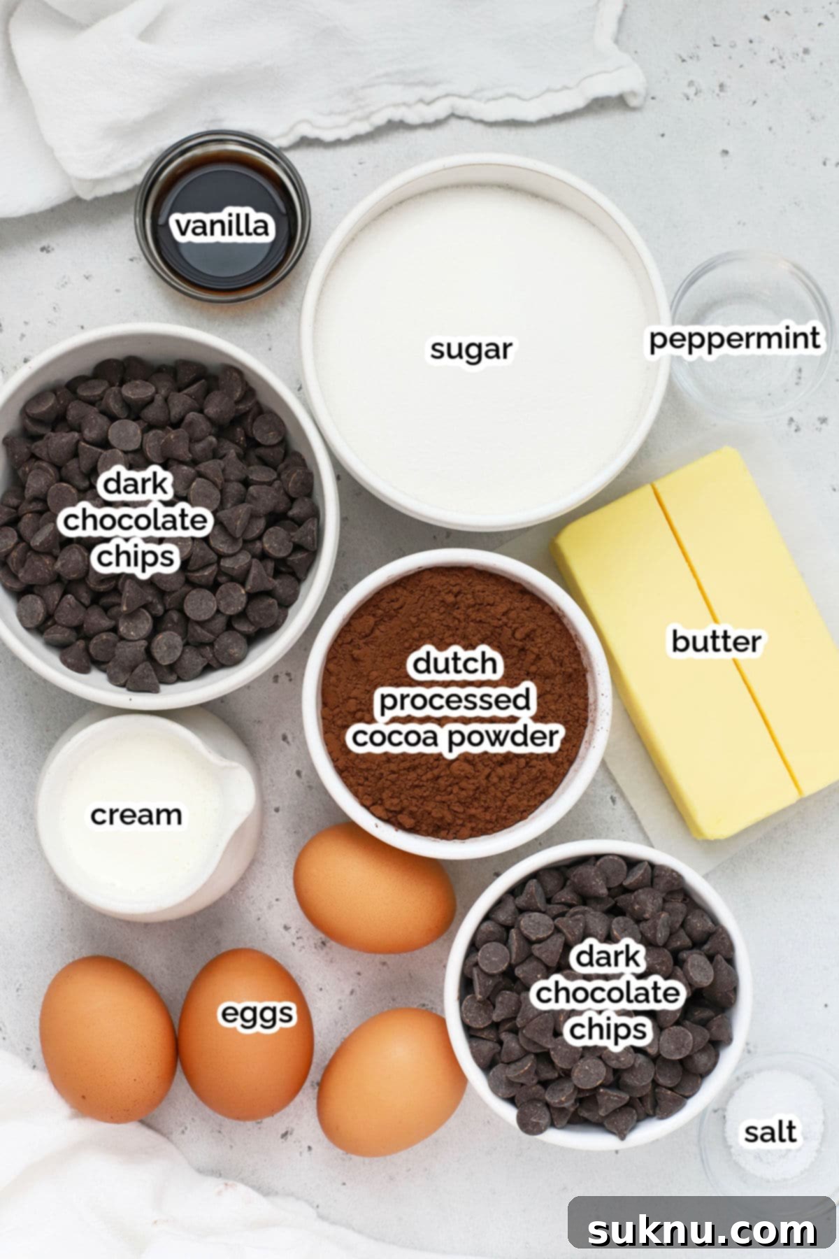 A collection of essential ingredients for making peppermint flourless chocolate cake, including sticks of butter, dark chocolate chips, granulated sugar, vanilla and peppermint extracts, and Dutch-processed cocoa powder.