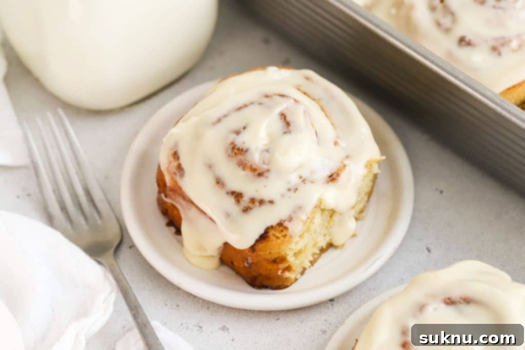 A beautifully frosted gluten-free cinnamon roll on a white plate, with a bottle of milk in the background.