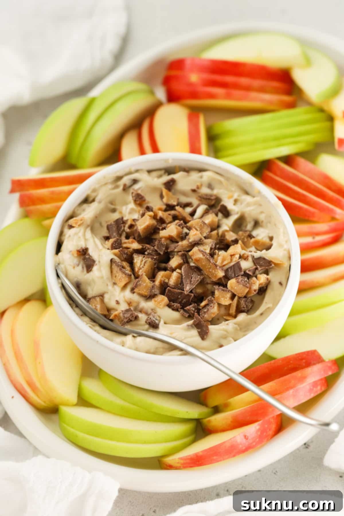 A beautifully presented bowl of creamy toffee apple dip, garnished with extra toffee bits, surrounded by vibrant red and green apple slices.