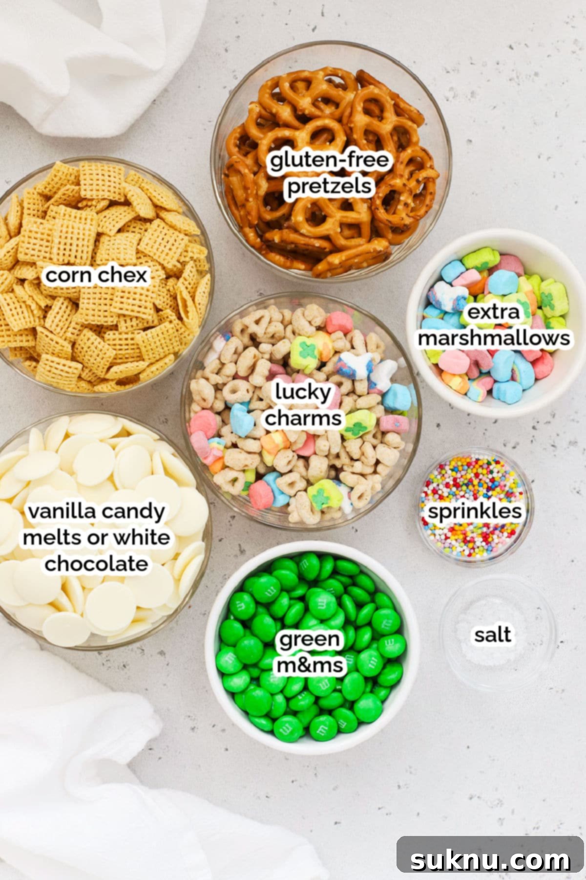 A flat lay photograph showcasing all the ingredients required for gluten-free leprechaun bait snack mix: corn Chex, Lucky Charms, gluten-free pretzels, green M&M's, white candy melts, sprinkles, and flaky sea salt.