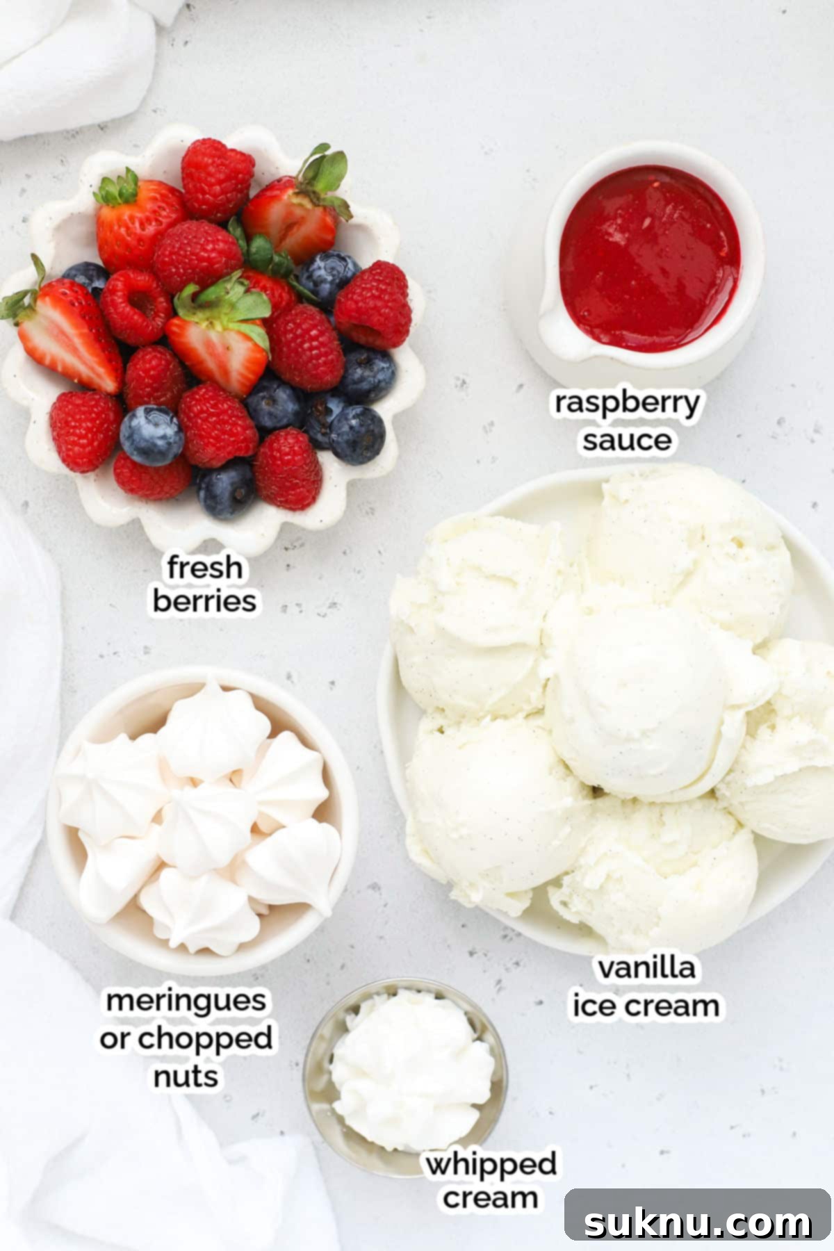 Ingredients laid out for making a Knickerbocker Glory sundae, including fresh raspberries, blueberries, strawberries, vanilla ice cream, meringue cookies, and coconut rolls
