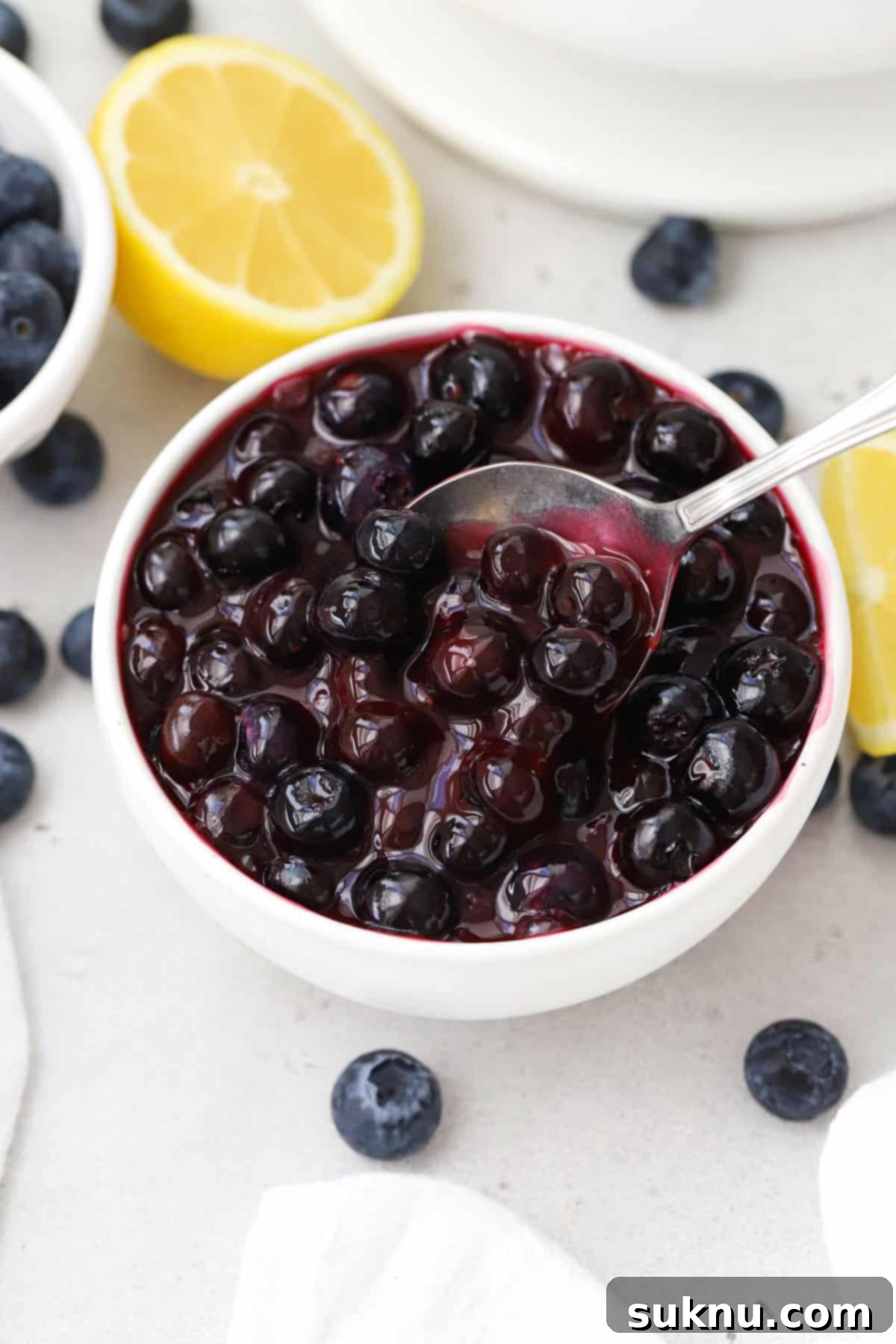 Easy Blueberry Sauce For Cheesecake And Beyond 6 A spoon dipping into a pristine bowl of fresh homemade blueberry sauce, showcasing its rich and inviting texture.