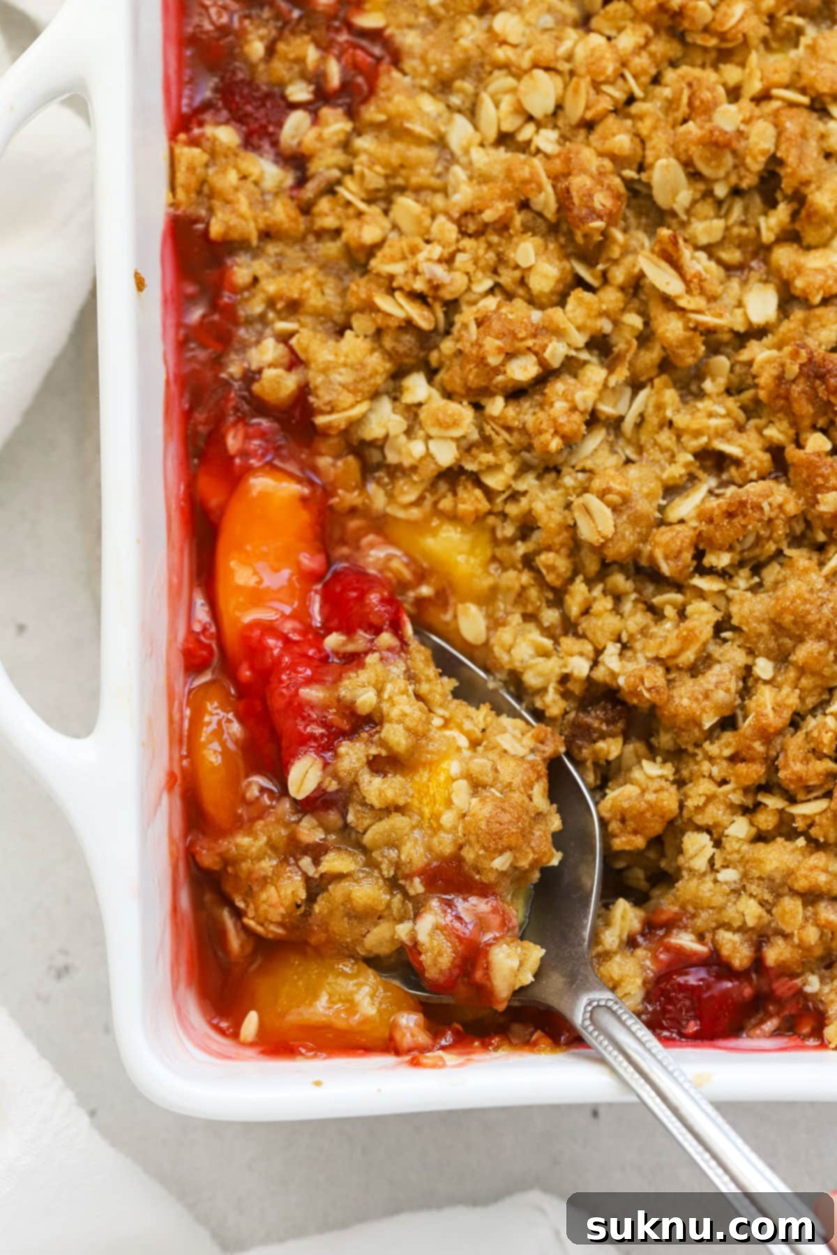 A close-up of a spoon scooping warm gluten-free raspberry peach crisp from a white baking dish, showing its delicious texture.