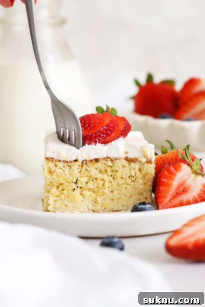 A close-up of a fork getting a bite out of a truly moist and luscious slice of gluten-free Tres Leches cake, beautifully soaked in three types of milk and crowned with delicate whipped cream.