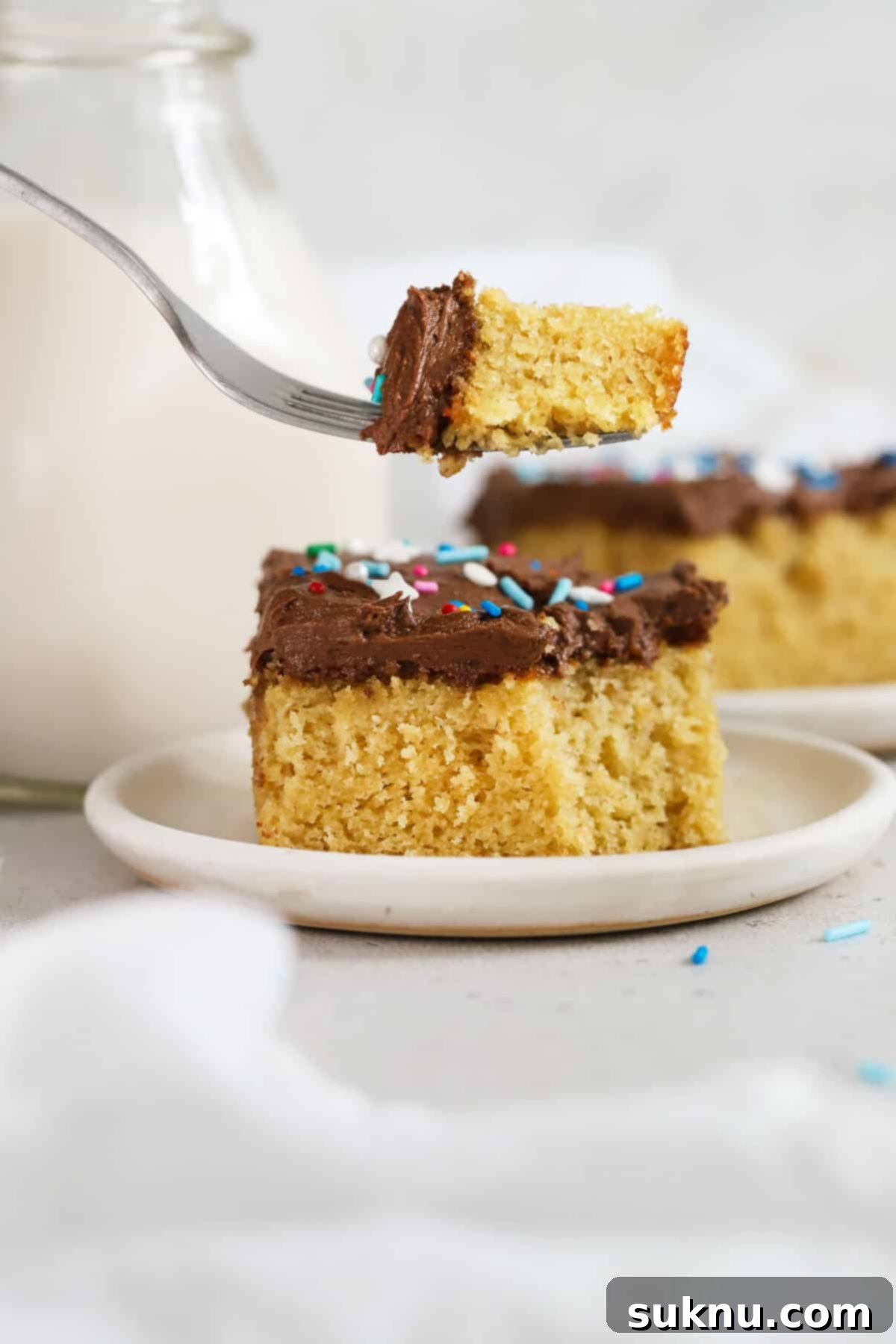 A close-up shot of a fork taking a bite out of a perfectly moist and fluffy slice of gluten-free yellow sheet cake with a generous layer of chocolate frosting.