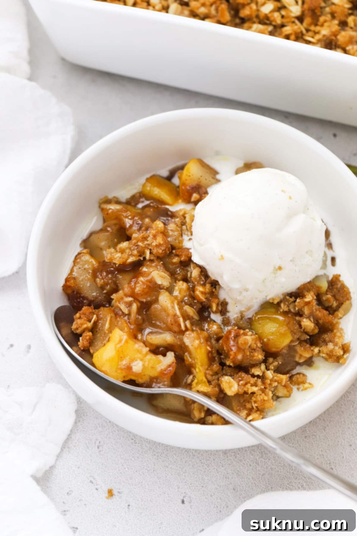 A spoon digging into a generous serving of gluten-free pear crisp, showcasing the tender fruit filling and golden oat topping, with melting vanilla ice cream.