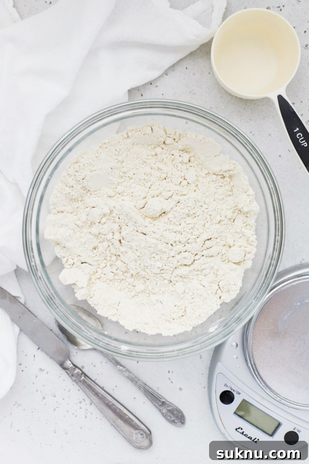 Master Flour Measurement For Perfect Baking 9 Overhead view of a bowl of flour with a measuring cup, spoon, knife, and scale, showcasing all tools for correct flour measurement