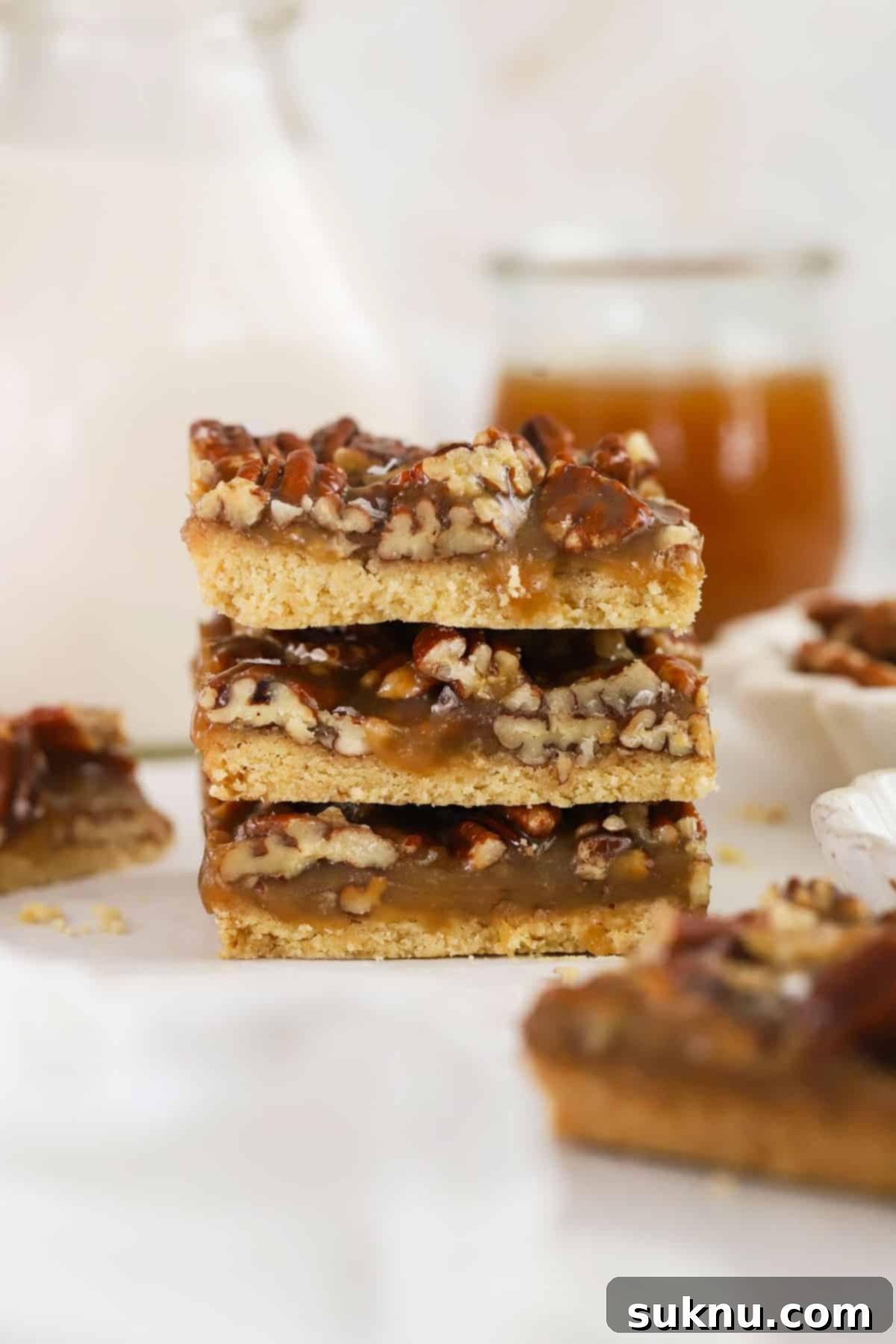 Sweet and Salty Gluten-Free Pecan Caramel Treats 9 Gluten-free Salted Caramel Pecan Bars stacked next to a bottle of milk and a jar of caramel sauce, creating a rustic scene.