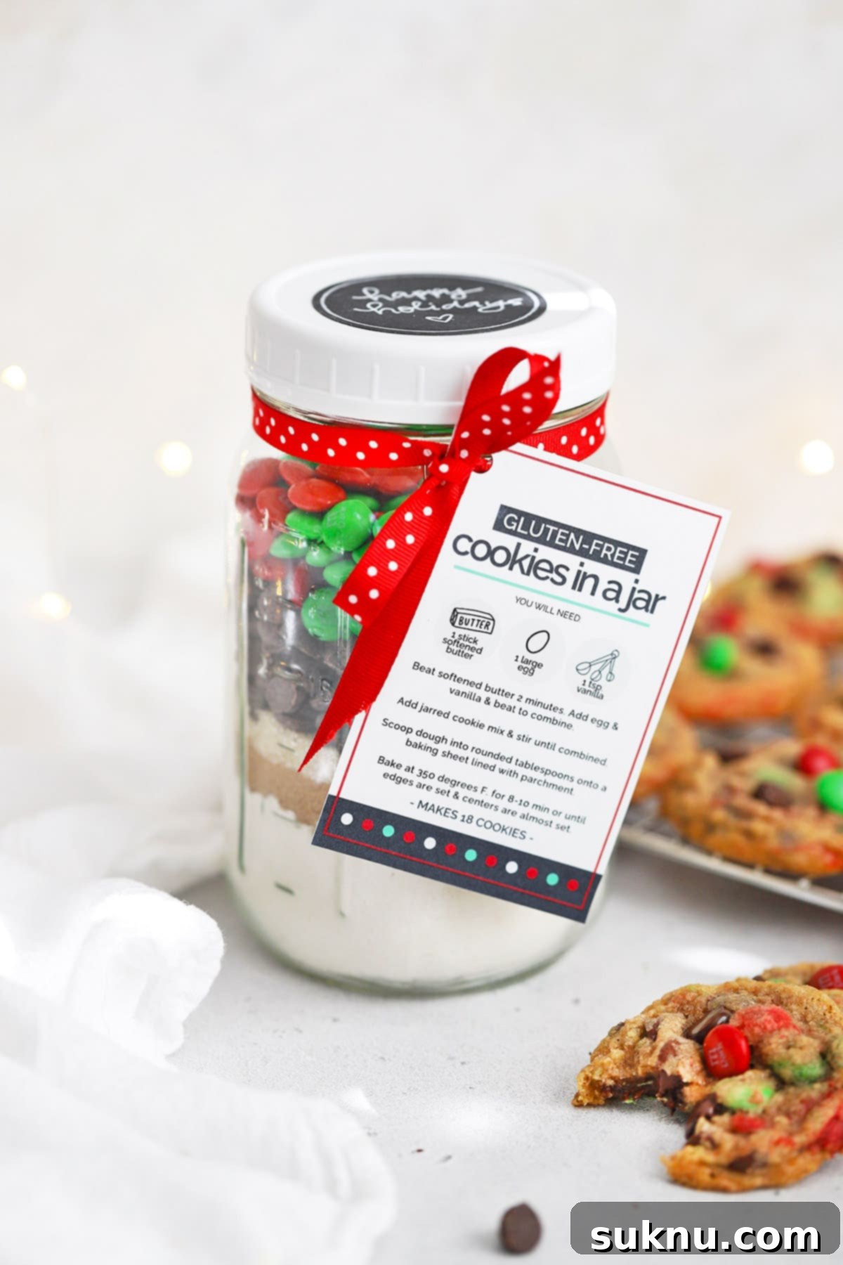 Beautifully layered gluten-free M&M cookie mix in a jar, presented with a free printable gift tag, against a backdrop of freshly baked gluten-free chocolate chip M&M cookies.