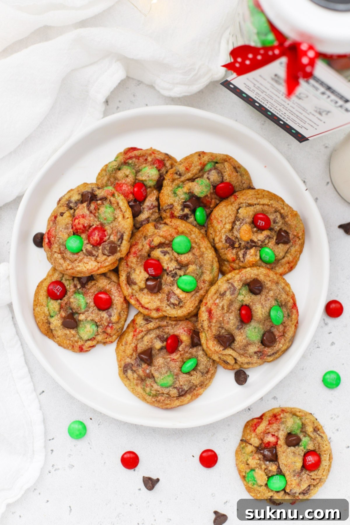 Appetizing overhead view of a plate of baked gluten-free M&M cookies, with a beautifully layered cookie mix in a jar subtly blurred in the background, showcasing the journey from mix to treat.
