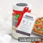 Beautifully layered gluten-free M&M cookie mix in a jar, presented with a free printable gift tag, against a backdrop of freshly baked gluten-free chocolate chip M&M cookies.