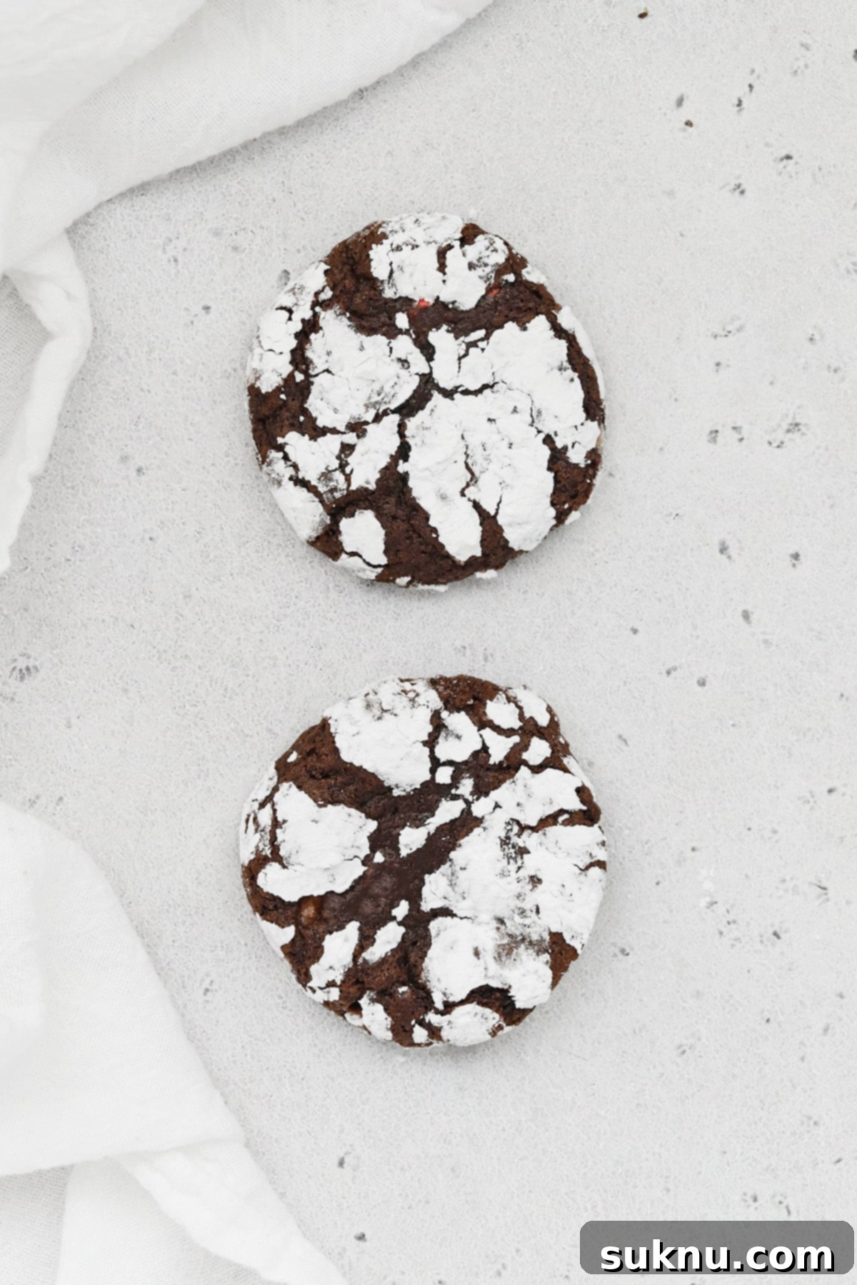 Two gluten-free peppermint chocolate crinkle cookies on a plain surface, highlighting their individual details and powdered sugar coating.