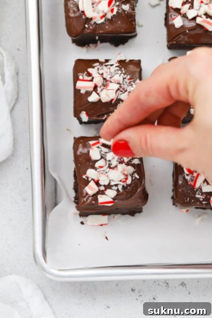 Crushed candy canes being generously sprinkled over a freshly chocolate-dipped gluten-free brownie bite on a baking sheet, creating a festive look.