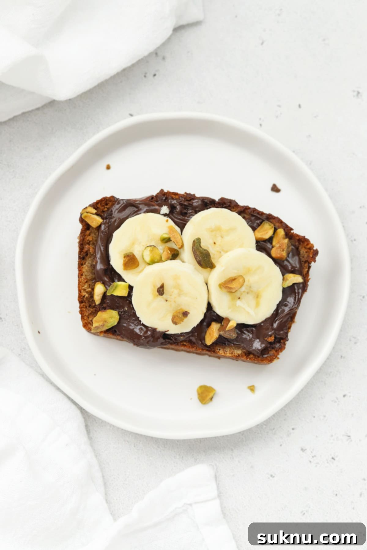 Slice of gluten-free banana bread topped with Nutella, fresh banana slices, and pistachios, presented on a white plate.
