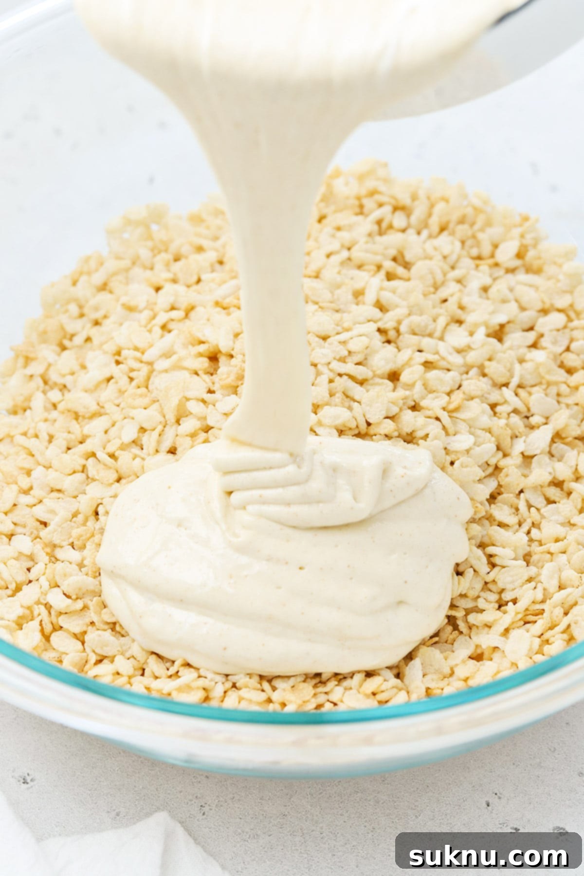 The melted brown butter and marshmallow mixture being poured over a bowl of gluten-free crispy rice cereal.