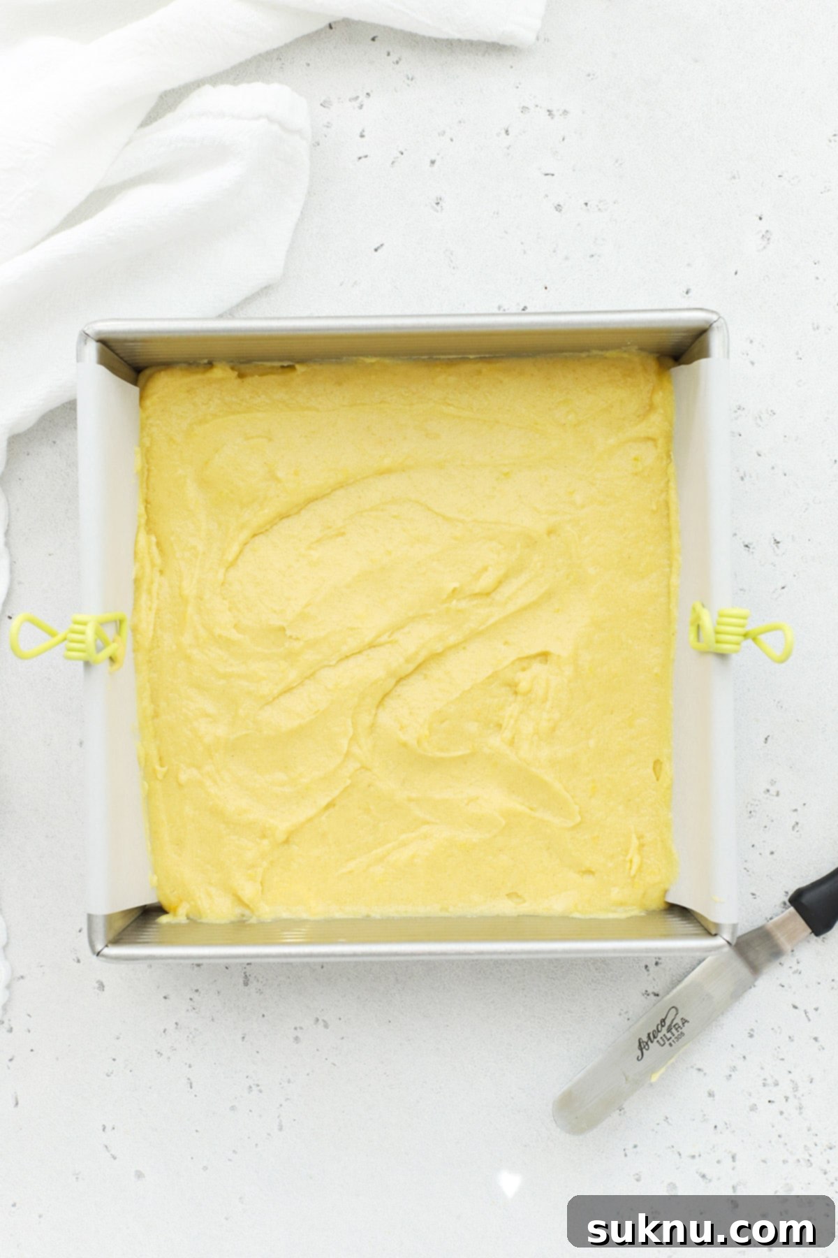 Smooth gluten-free lemon brownie batter being spread evenly into a parchment-lined baking pan with an offset spatula.