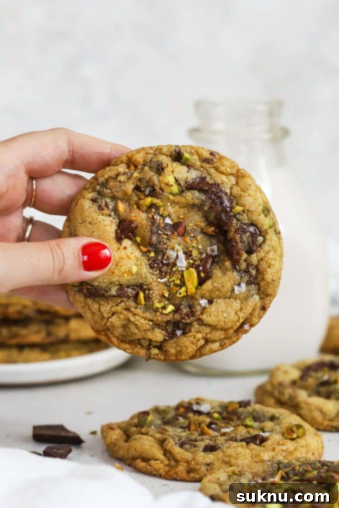 A hand holding a beautifully baked gluten-free brown butter chocolate chunk pistachio cookie, showcasing its rustic appearance and delightful ingredients.