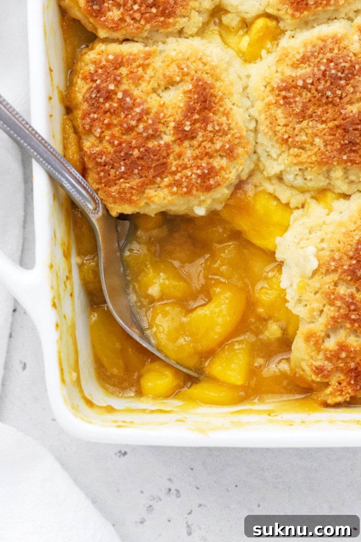 Simple Delicious Gluten-Free Peach Cobbler 3 A serving spoon gracefully scooping into a warm, inviting gluten-free peach cobbler, revealing the tender peaches and golden biscuit topping beneath the crisp crust.