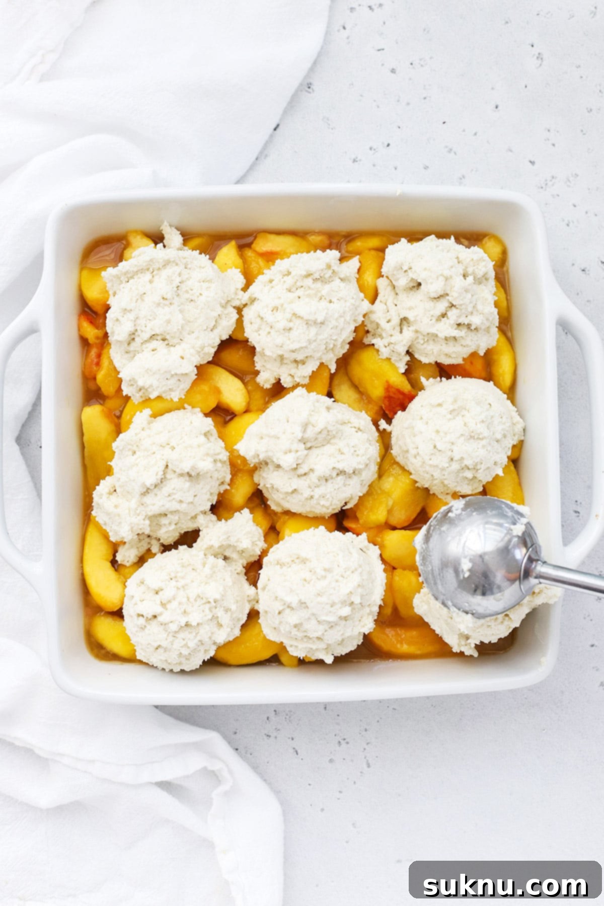 Simple Delicious Gluten-Free Peach Cobbler 8 A large cookie scoop carefully placing sweet gluten-free biscuit dough mounds over the glistening peach filling in a baking dish, creating the cobbler's top layer.