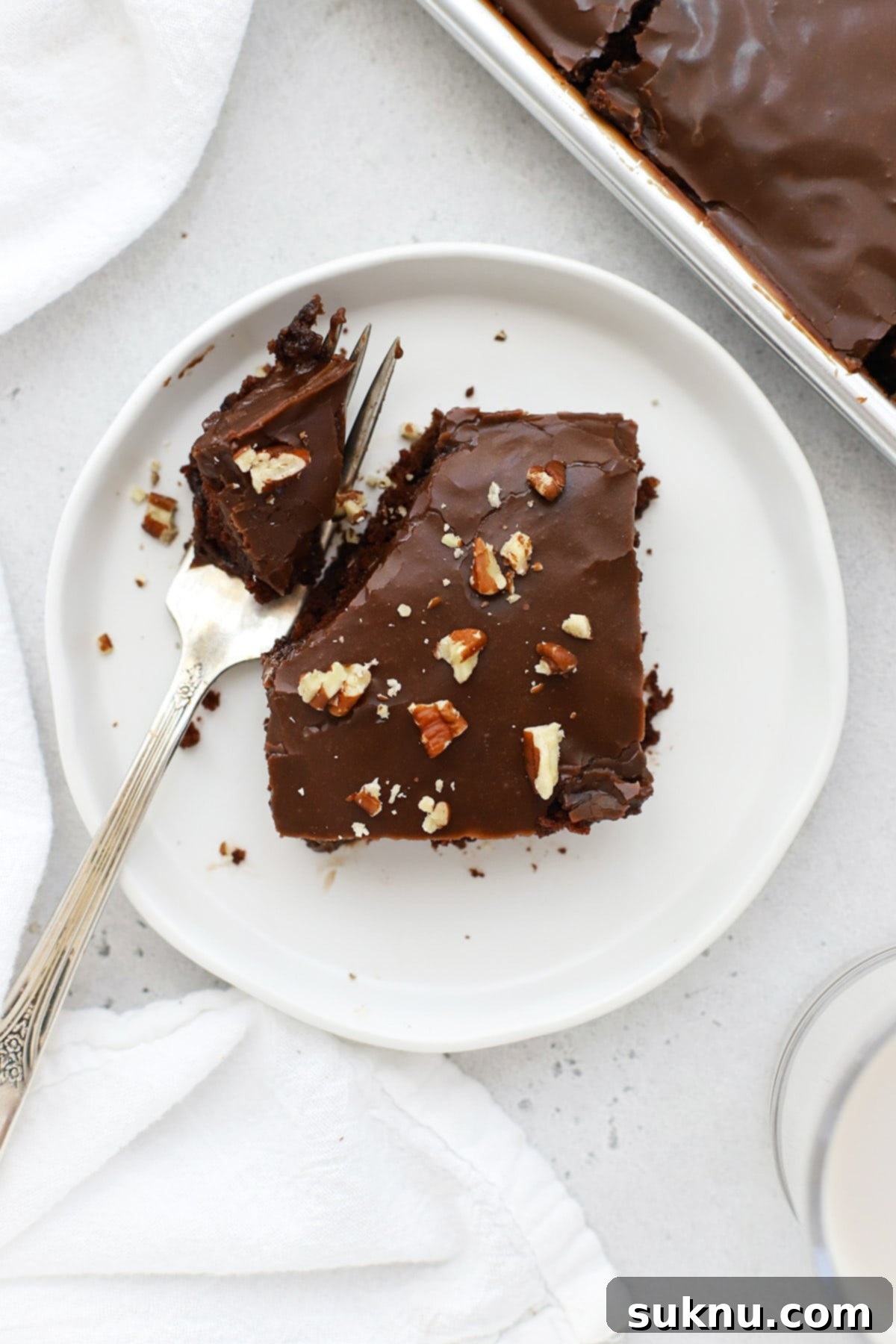 A slice of gluten-free Texas chocolate sheet cake generously topped with toasted chopped pecans, sitting on a white plate.