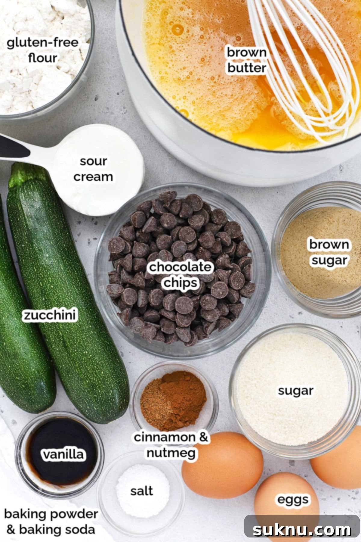 A collection of ingredients laid out for making gluten-free zucchini muffins, including flour, eggs, butter, zucchini, and chocolate chips.