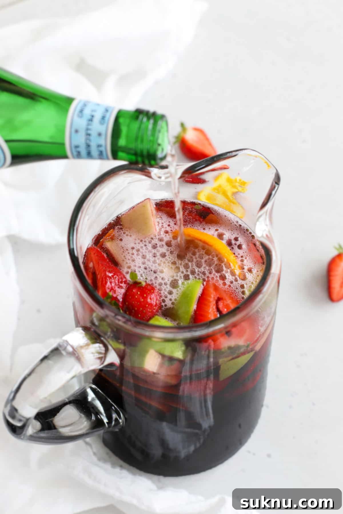 A person pouring sparkling water into a pitcher of non-alcoholic sangria, already filled with juices and fruit