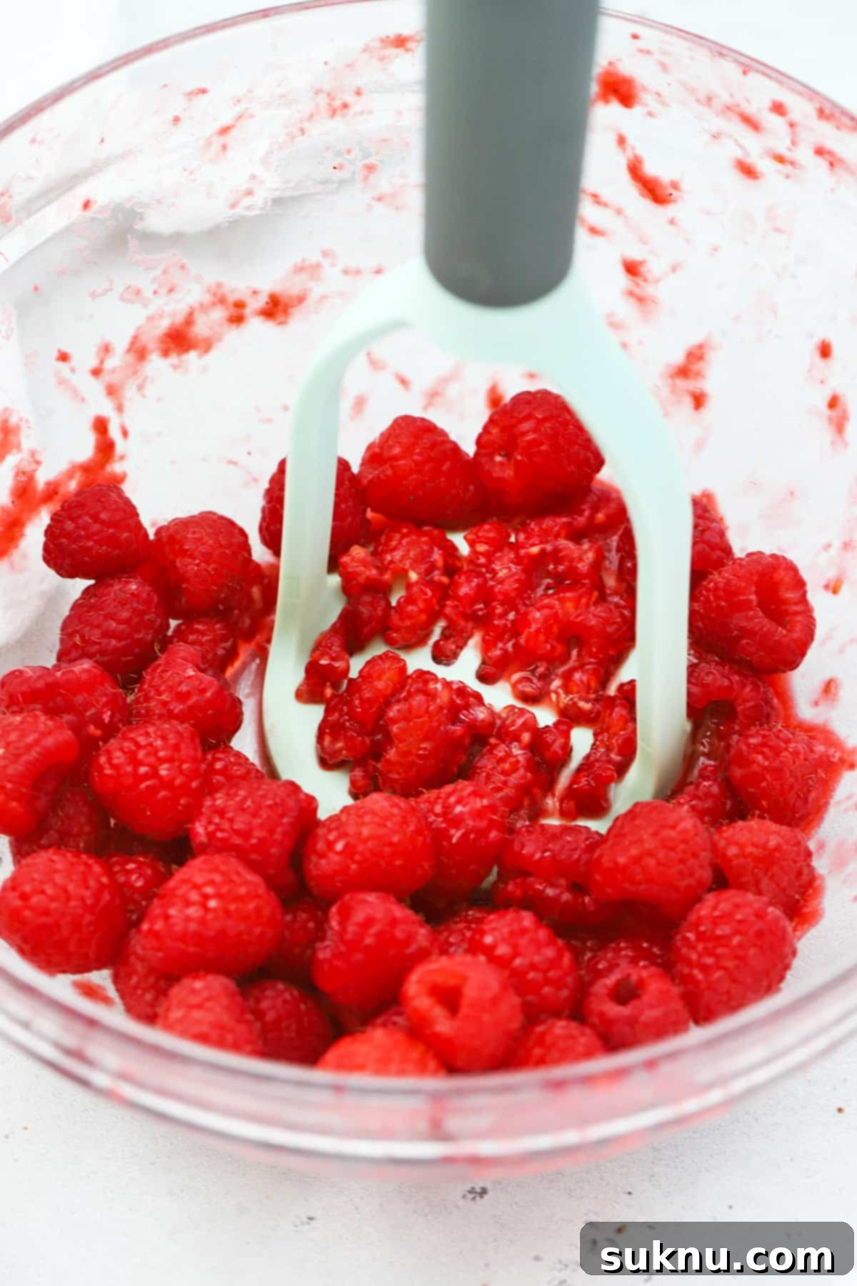 Mashing fresh raspberries in a bowl with a potato masher, preparing them for homemade freezer jam.