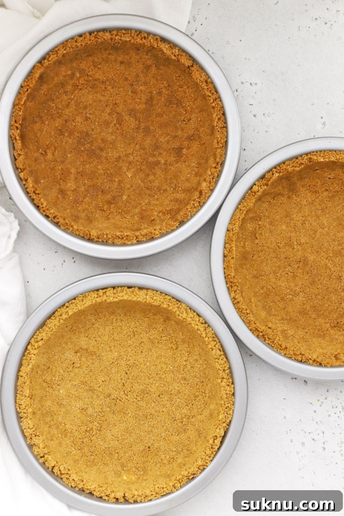 Three perfectly formed gluten-free graham cracker crusts nestled in individual light-colored metal pie pans.
