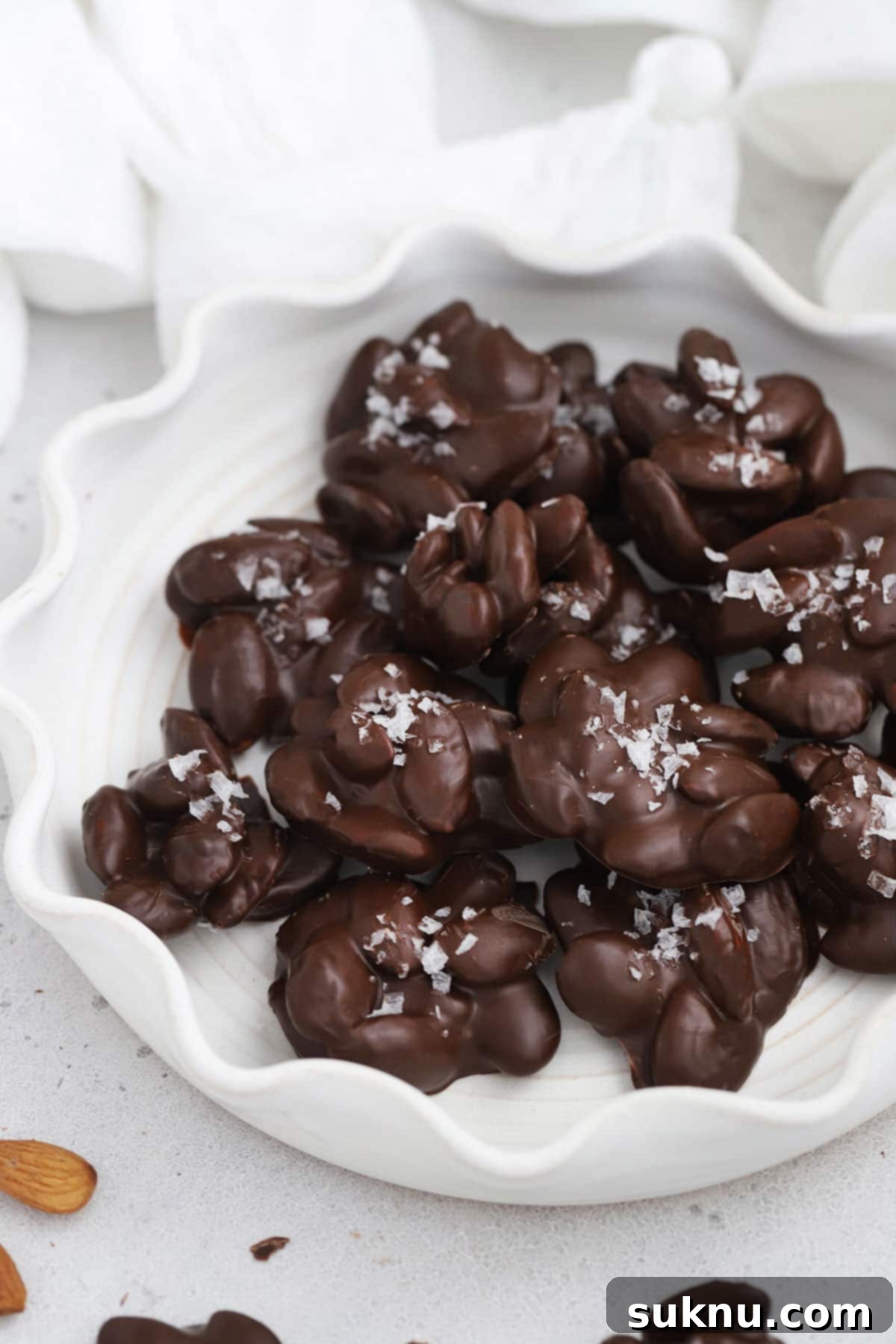 Front view of a ruffled white plate of dark chocolate almond clusters topped with flaky sea salt, highlighting their crunchy texture and gourmet appeal