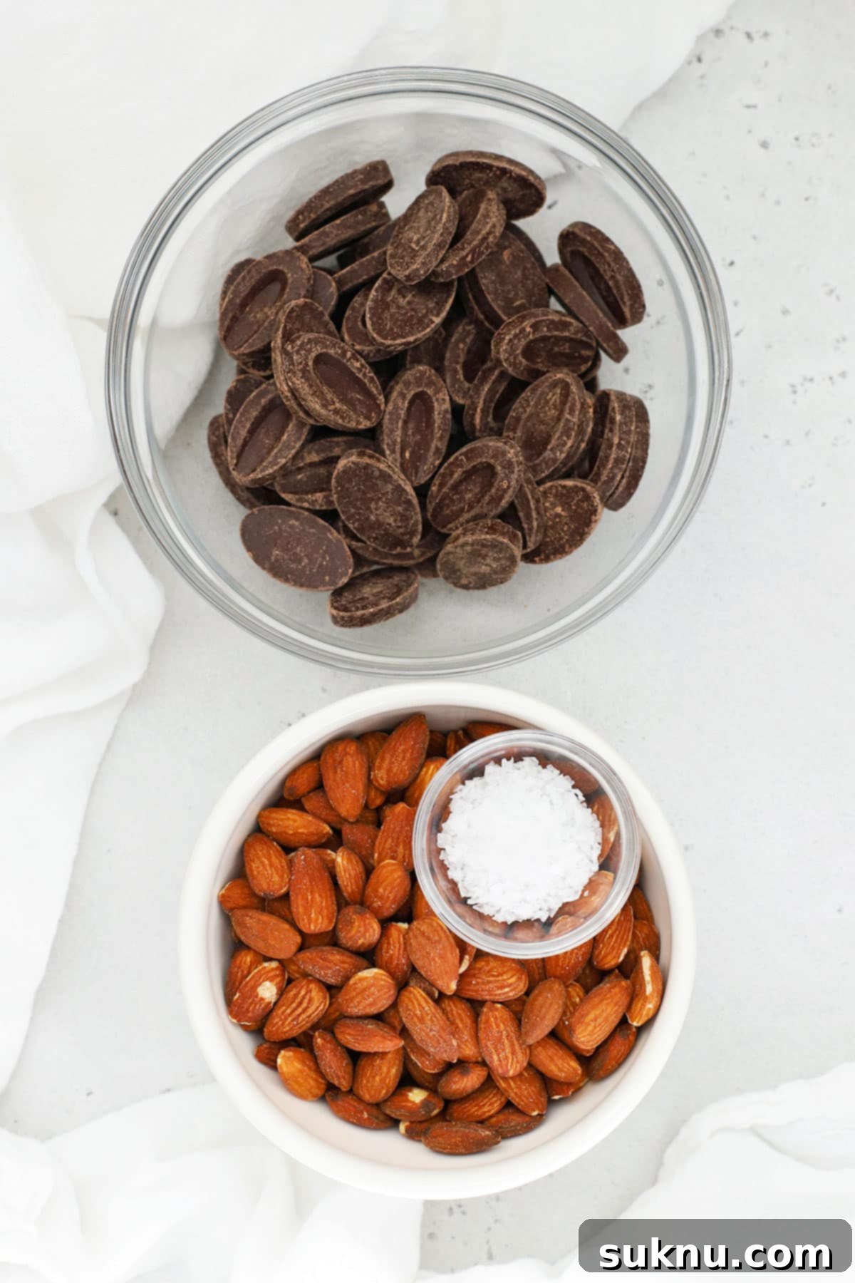 Overhead view of ingredients for dark chocolate almond clusters, including chocolate, almonds, and flaky sea salt, laid out in small bowls
