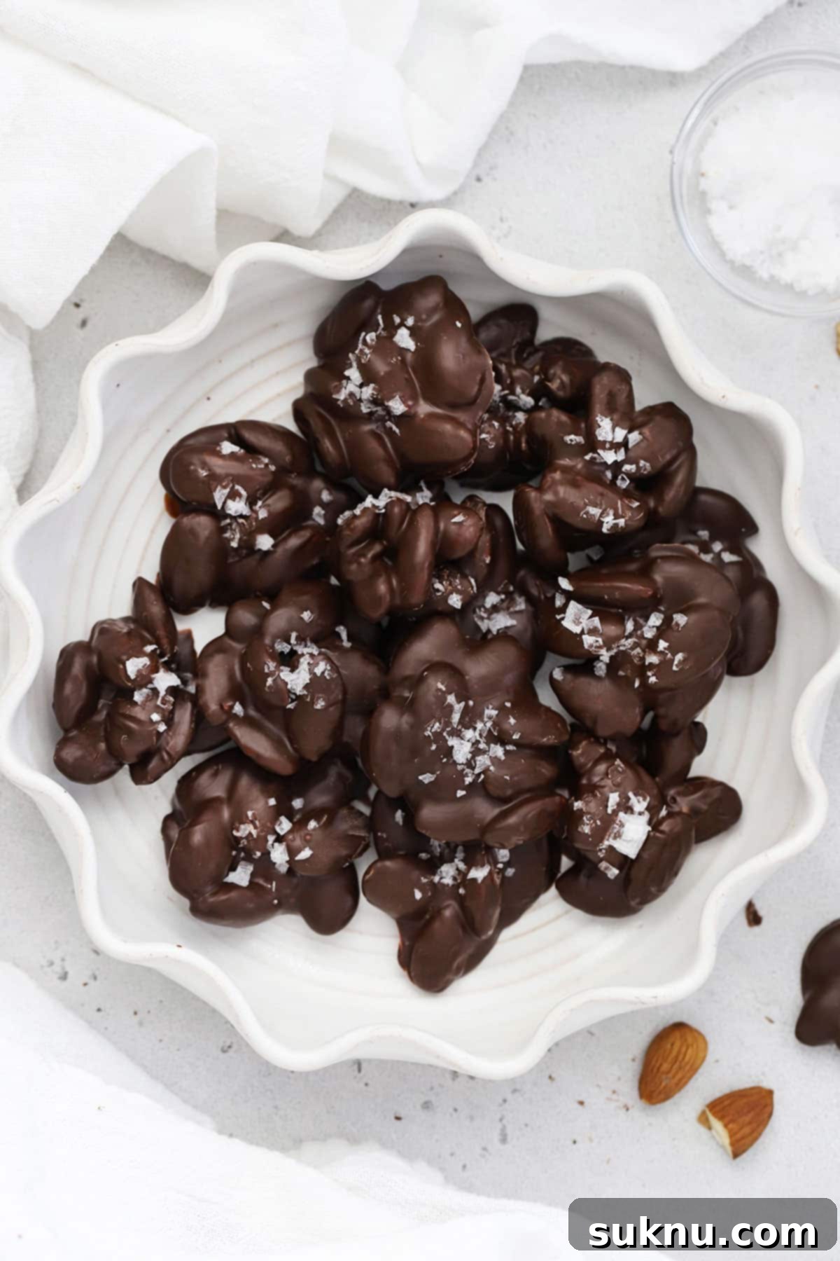 Dark chocolate almond clusters with flaky salt arranged beautifully on a white ruffled plate, ready to be served