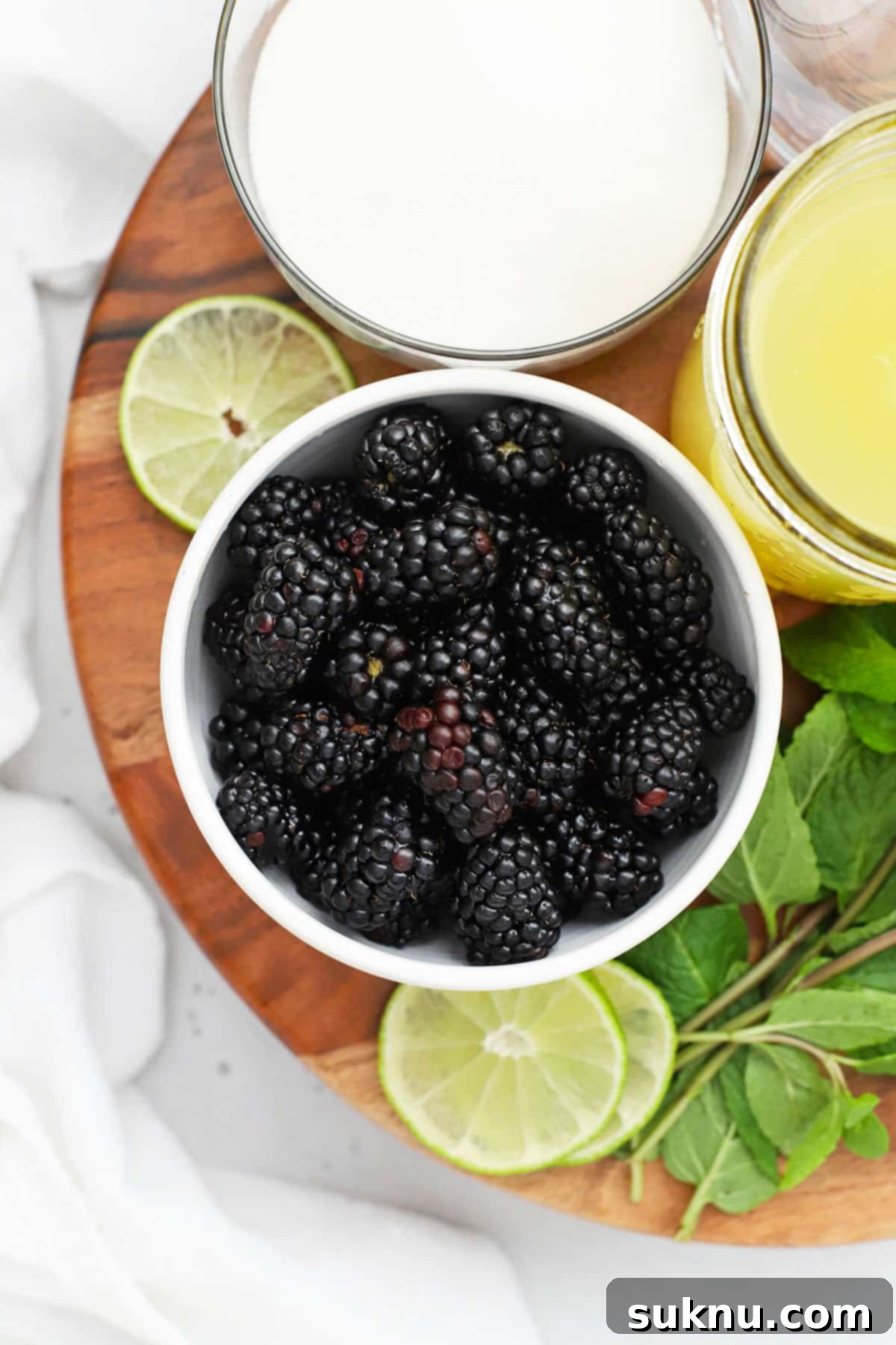 Close-up of fresh ingredients for blackberry limeade including limes, blackberries, mint, and a pitcher of simple syrup.