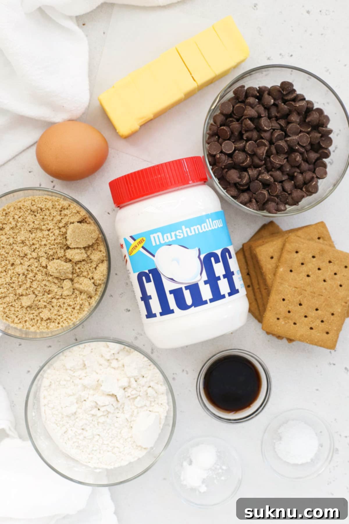 Ingredients for gluten-free s'mores bars laid out on a white background, including butter, brown sugar, flour, graham cracker crumbs, marshmallow fluff, and chocolate chips.