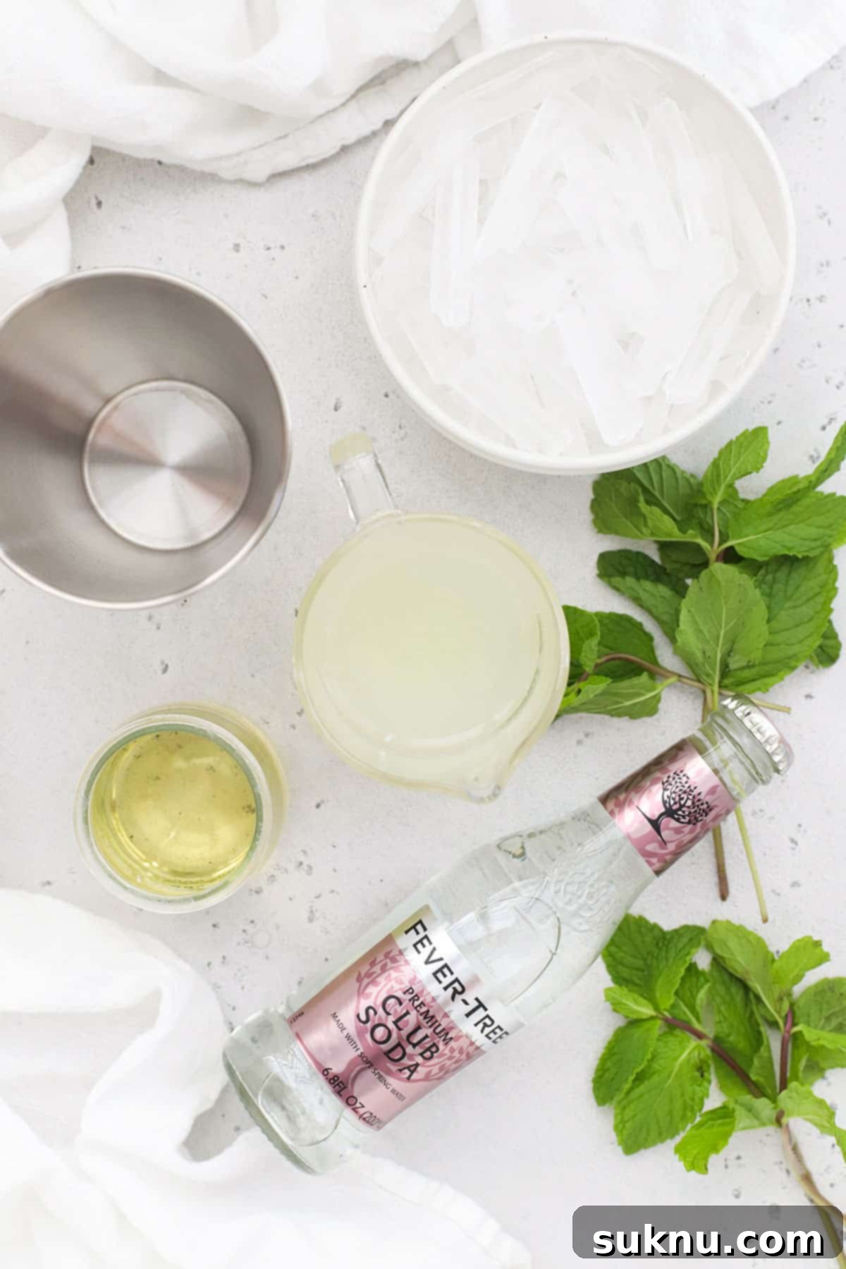 Disneyland Style Mint Julep 4 Individual ingredients for a virgin mint julep mocktail are artfully arranged: a pitcher of homemade mint simple syrup, a bowl of fresh mint leaves, a glass filled with lemonade, and a bottle of lemon-lime soda, all set against a light background, highlighting their freshness and vibrancy.