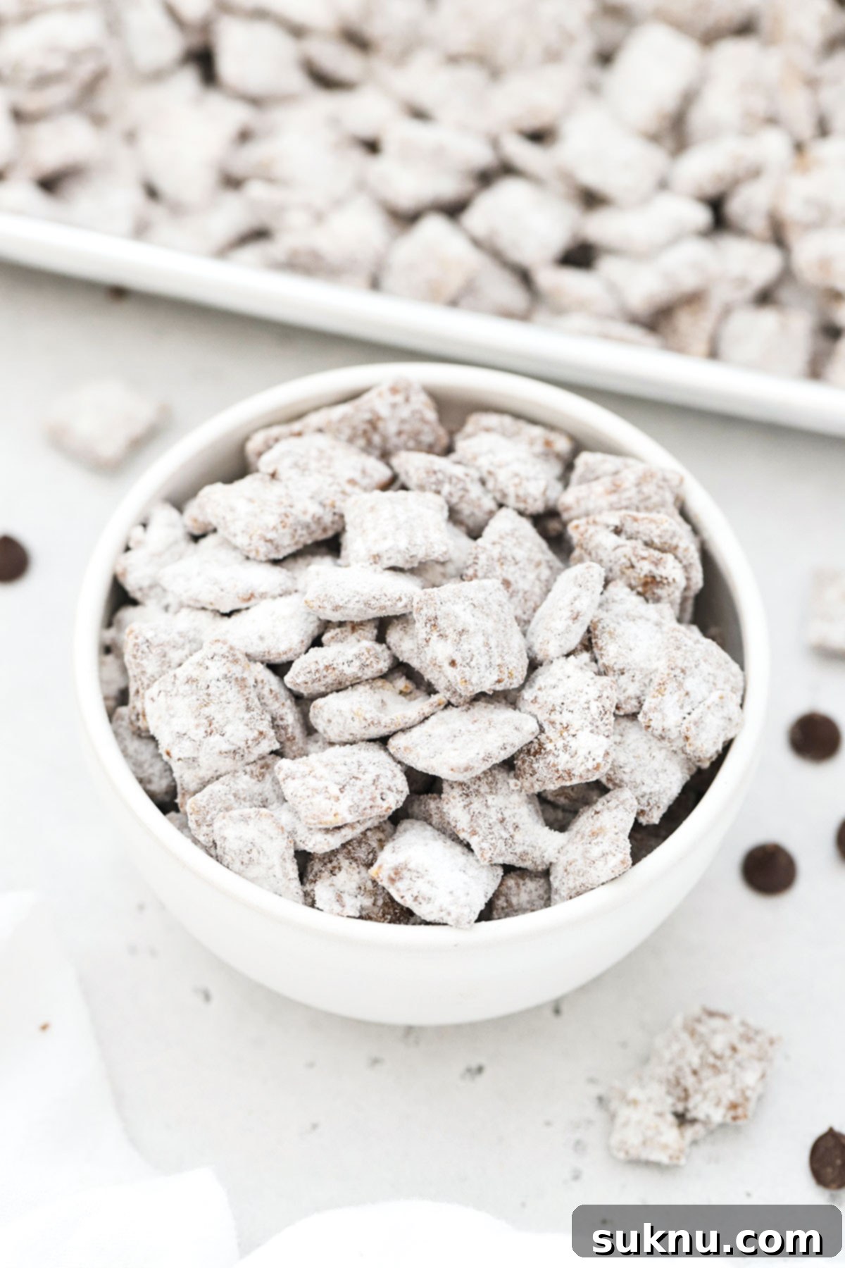 A close-up of gluten-free puppy chow in a white bowl, showcasing the distinct powdered sugar coating and clusters.