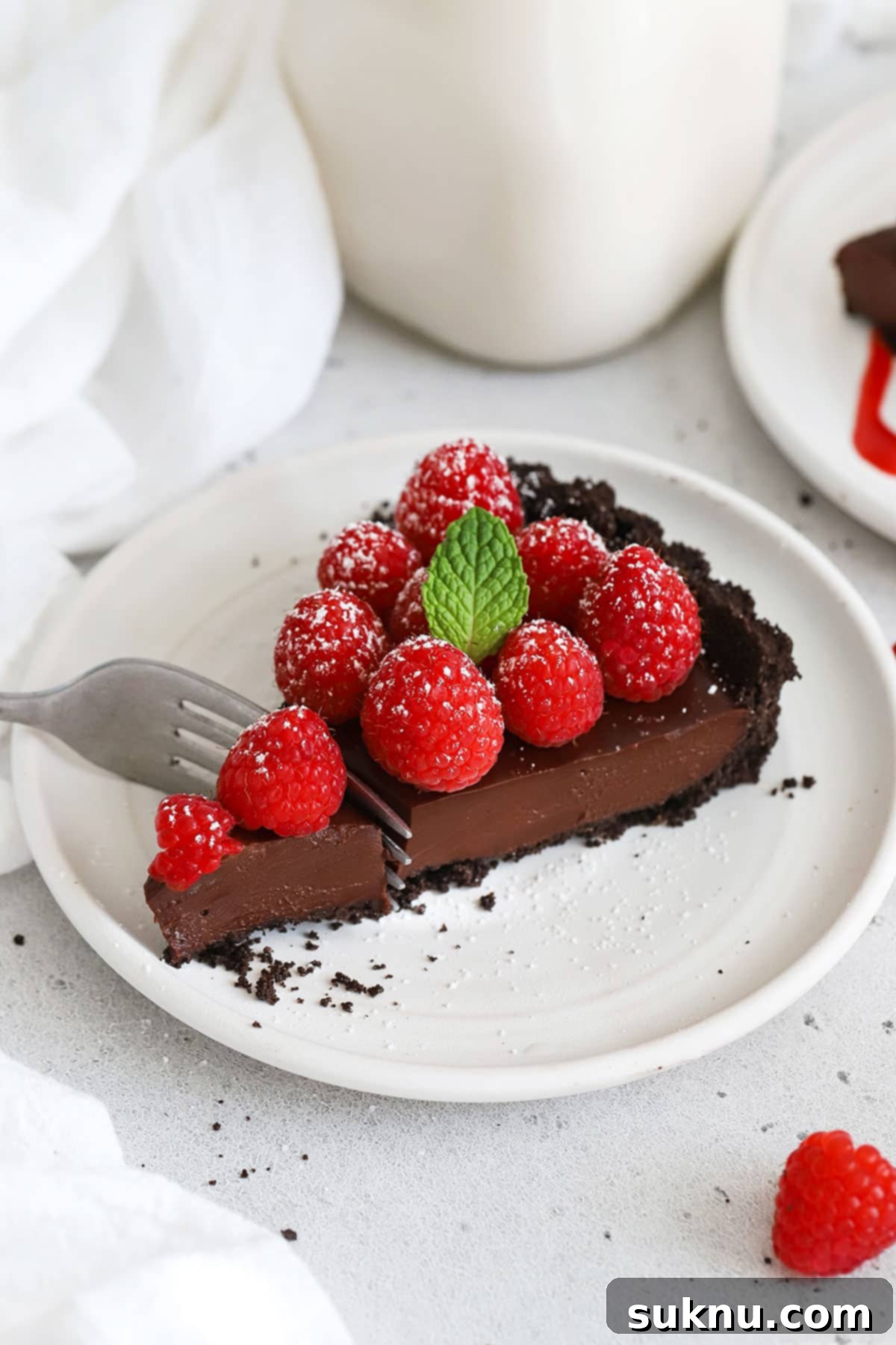 A close-up of a single slice of gluten-free chocolate raspberry tart on a white plate, highlighting its smooth ganache and crumbly crust.