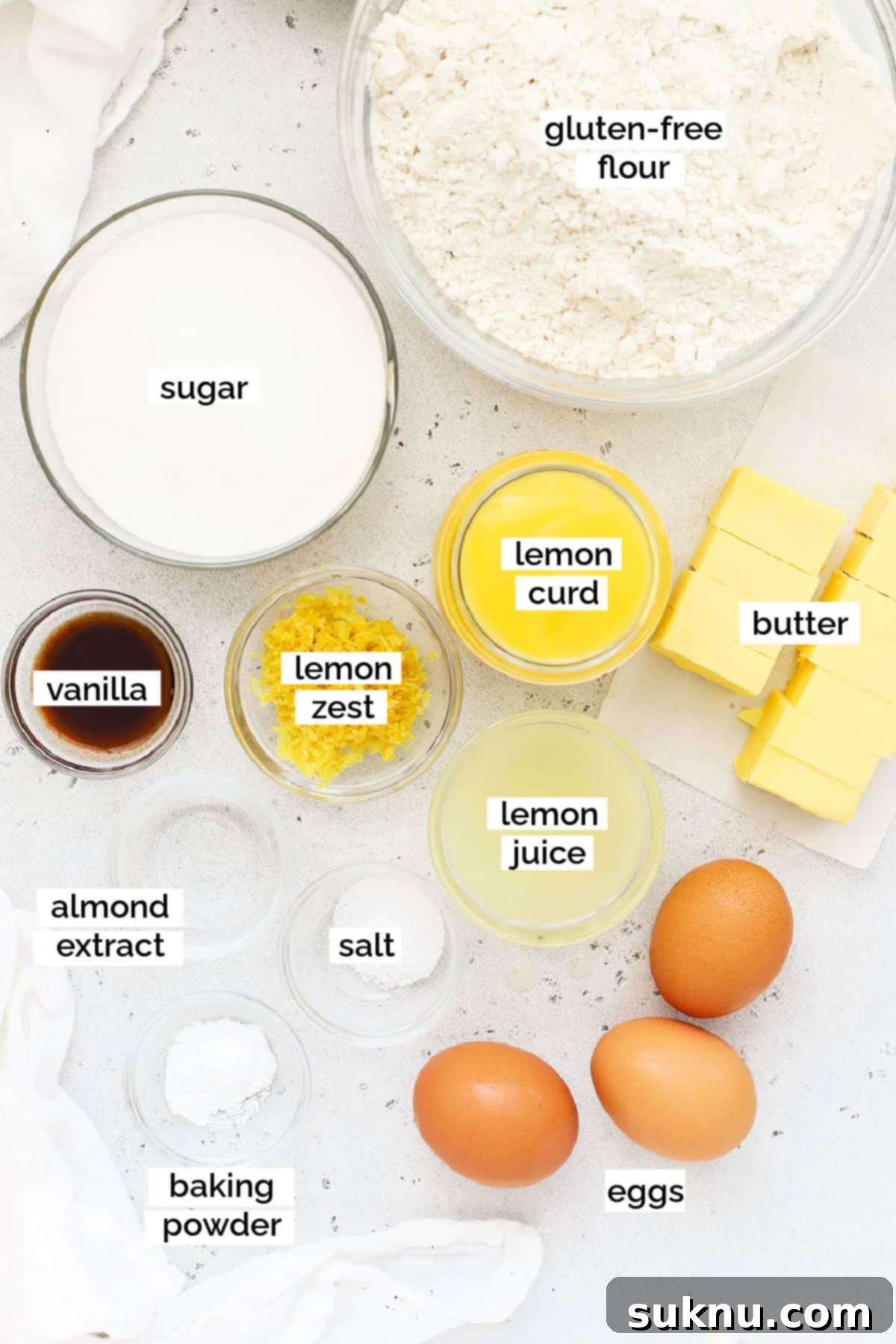 Zesty Gluten-Free Lemon Curd Gems 3 A collection of ingredients for gluten-free lemon curd cookies laid out on a white surface, including lemons, flour, sugar, butter, and eggs.